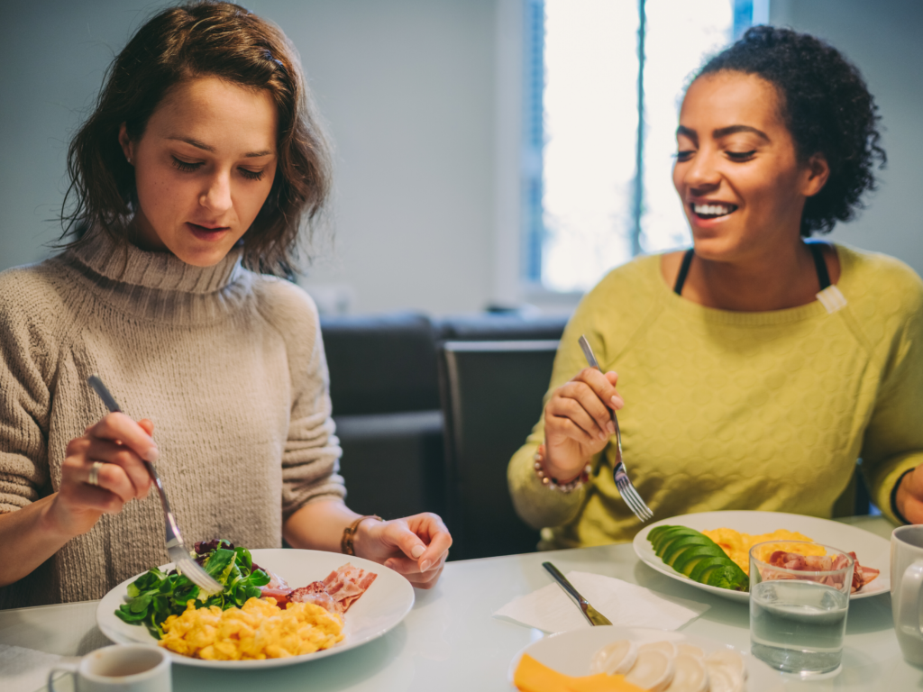 Two women sit down to eat together. Woman on the left has pale skin and is looking, with concentration, at a plate filled with meats, salad, and cheesy pasta. Woman on the right is watch the woman on the left with a happy expression, as if she were mid-conversation and beginning to laugh. 