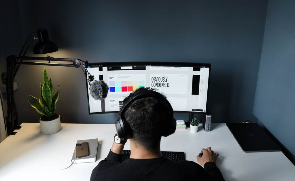 Decorative image. Freelance graphic designer sits at a computer screen with graphic design software on the screen. He wears a pair of headphones and is facing away from the camera. 