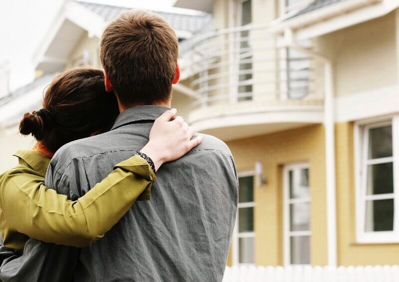 Decorative Image. Man and woman stand with their backs to the camera, hugging and looking at the exterior of a house.