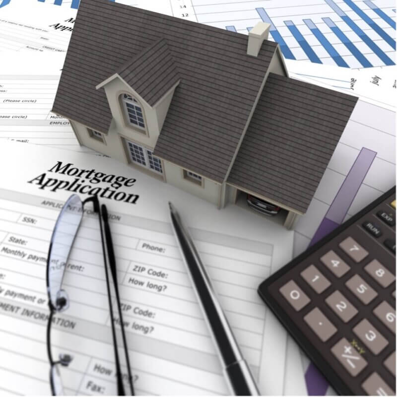 Decorative Image. A small toy house, pen, pair of glasses, and a calculator sit on a surface covered in documents and forms. The document in focus sits under the house, pen, and eye glasses and says Mortgage Application